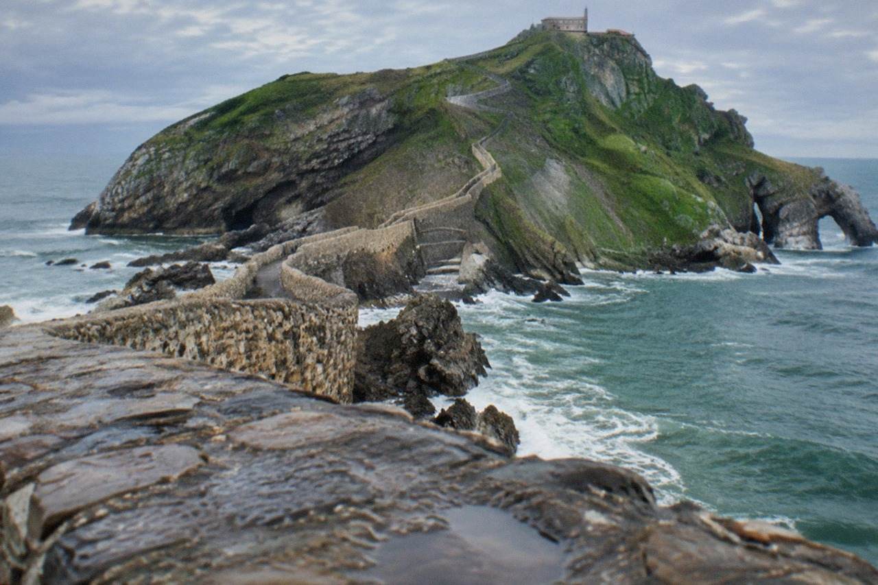A rocky islet on the Basque coast with a stone causeway and a long staircase climbing to a small hermitage at the top