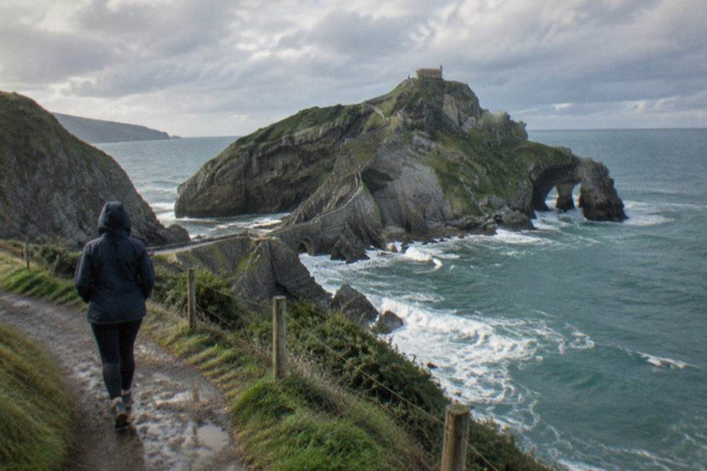 A visitor in a rain jacket looking out at the Gaztelugatxe causeway from the clifftop path, waves breaking against the rocks below