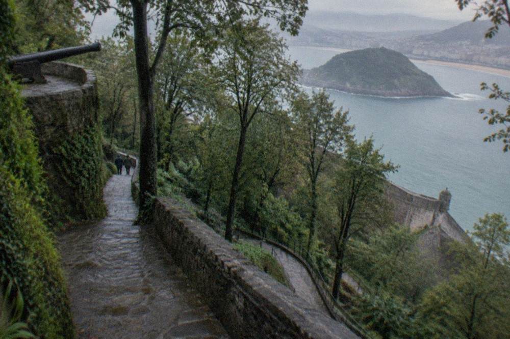 A stone path winding uphill through trees on Monte Urgull toward the fortress walls above the old town