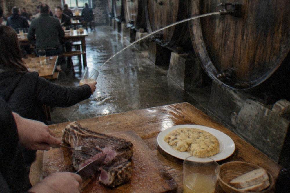 A thick txuleta rib steak being cut on a wooden board at a Basque cider house table