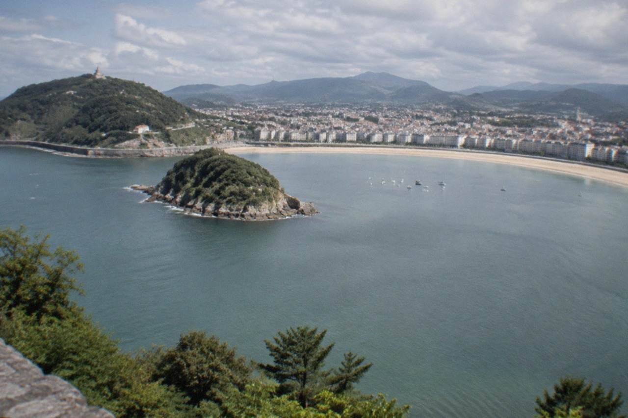 The curved beachfront of La Concha bay in San Sebastián seen from above, with the island and green hills behind the water