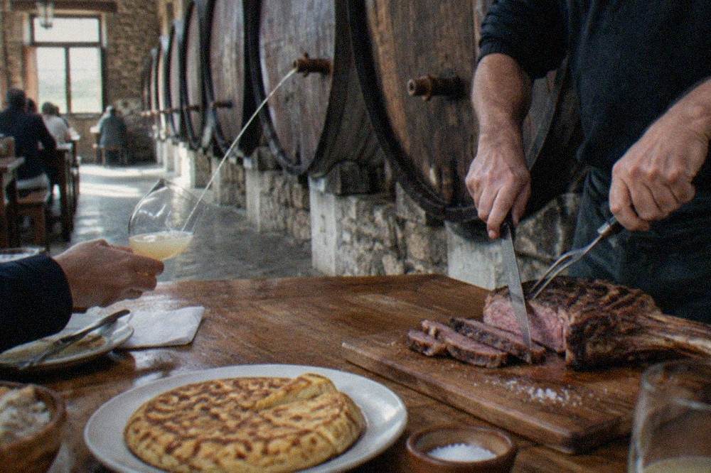 A large grilled rib steak being carved at a wooden table in a Basque cider house