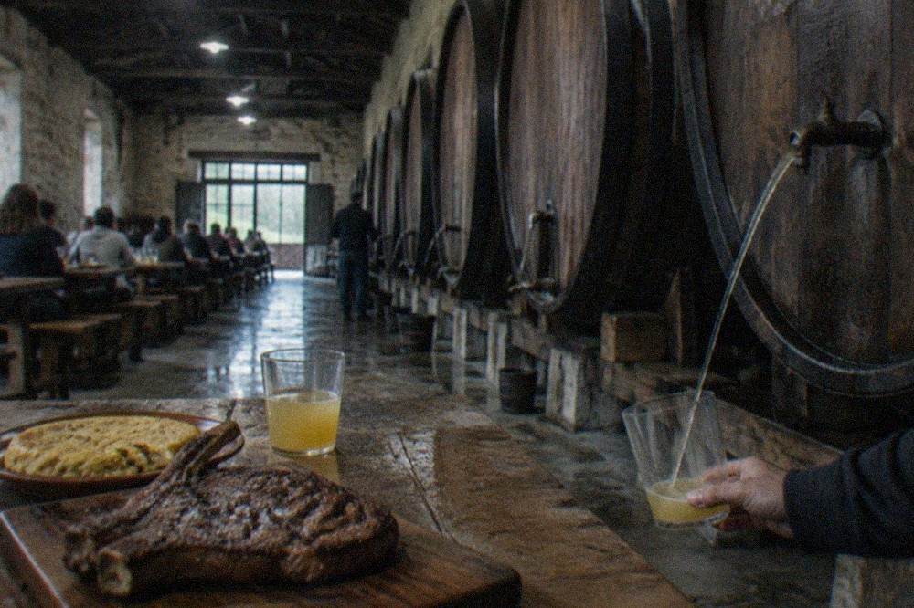 A grilled txuleta steak on a wooden board beside glasses of Basque cider, on a rustic cider house table.