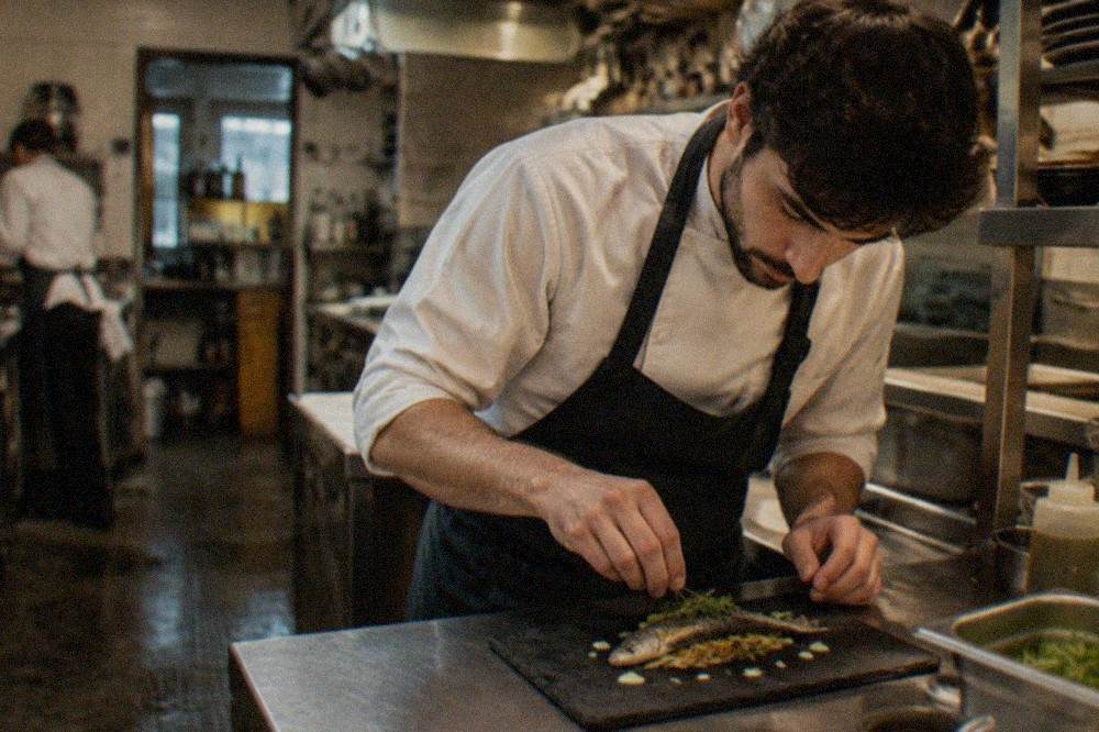 A chef plating a small fish course on a slate plate in a small restaurant kitchen in the Basque Country