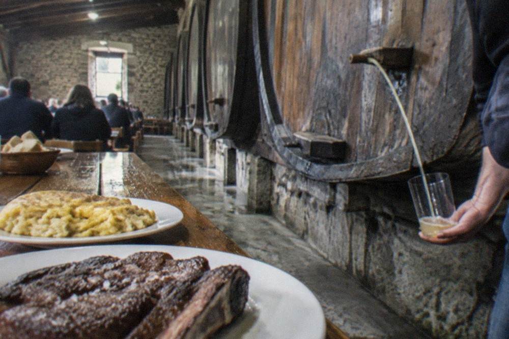 A stream of cider pouring from a barrel into a glass held at waist height in a Basque cider house