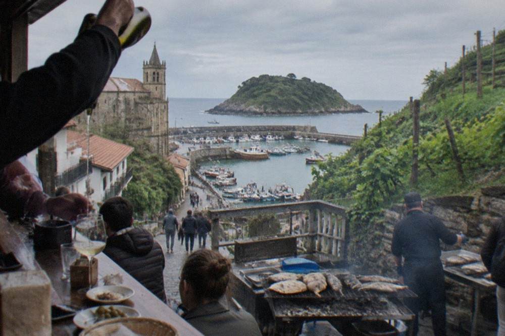A waiter pouring txakoli from high above into a glass on a busy pintxo bar counter