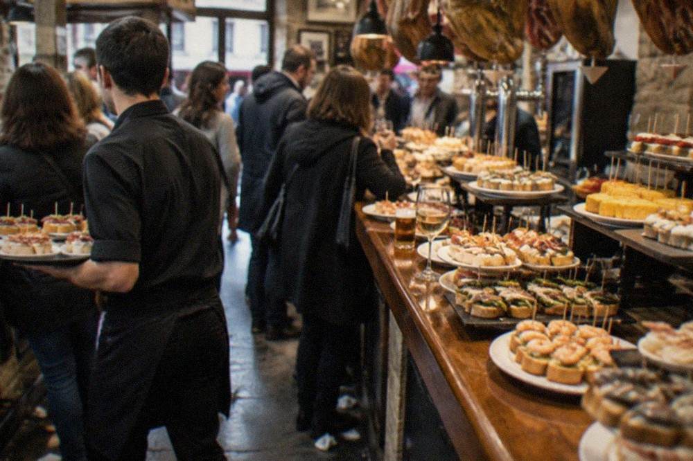 Waiter carrying small plates on a tray through a busy pintxo bar in the evening
