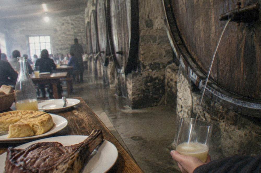 A cider barrel with a thin stream of cider pouring into a held-low glass at a Basque cider house