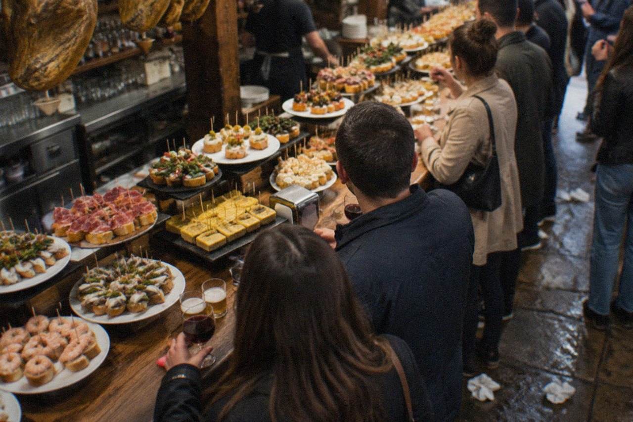 Evening crowd standing at a pintxo bar counter in the old town of San Sebastián, with plates of small skewered snacks on the bar top