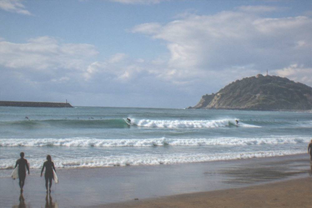 Waves breaking at Zurriola beach with surfers in the water and Monte Ulia in the distance