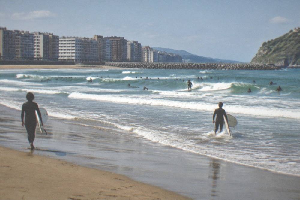Zurriola beach in San Sebastián with surfers in the waves and the Gros apartment blocks behind the sand
