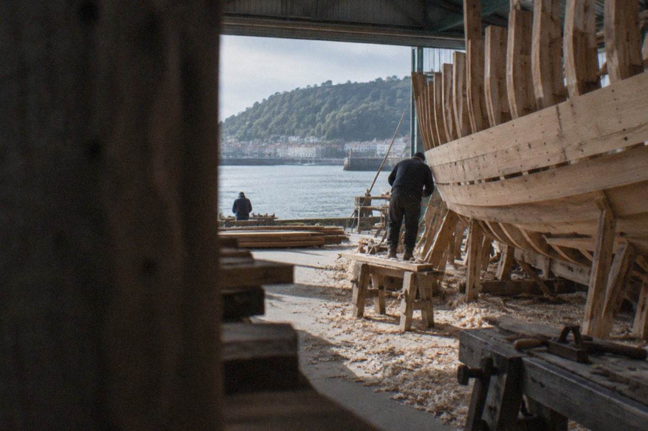 A wooden ship hull under construction inside a warehouse workshop by the sea