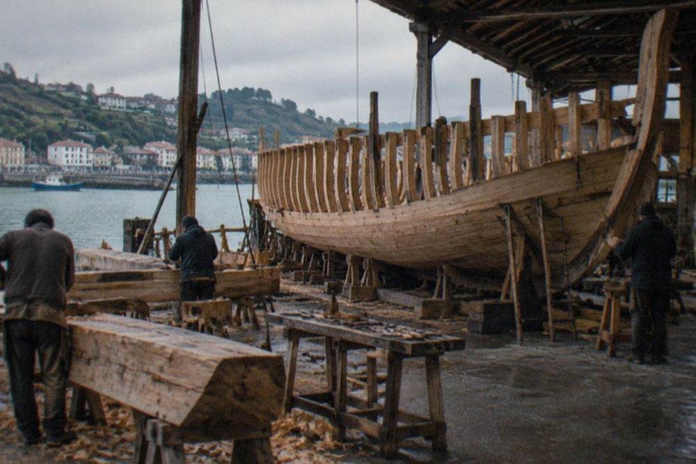 Carpenters shaping large wooden beams with hand tools in an open workshop