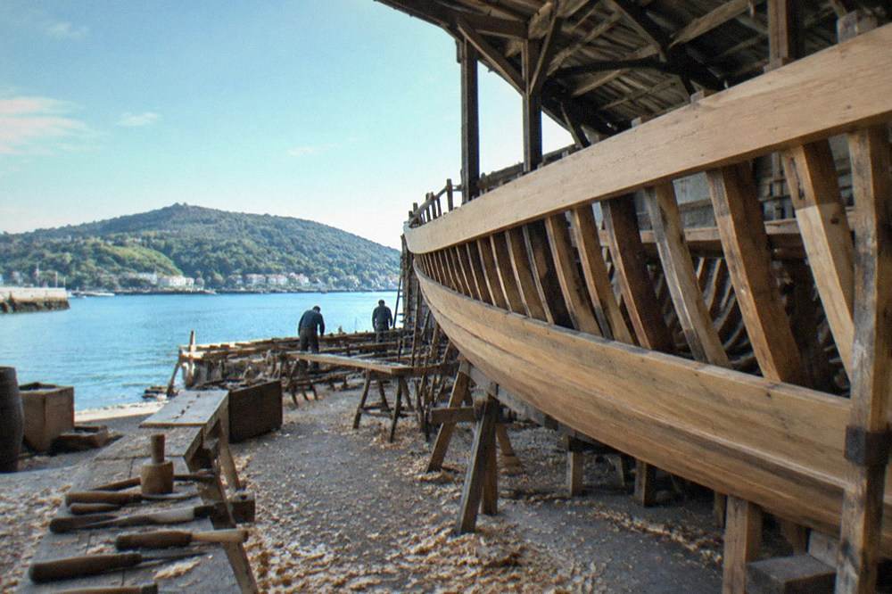 A wooden sailing ship hull with ribs and planks visible inside a large shed