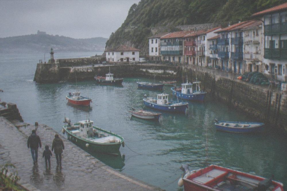 A family walking along a harbour wall with fishing boats moored in a narrow bay