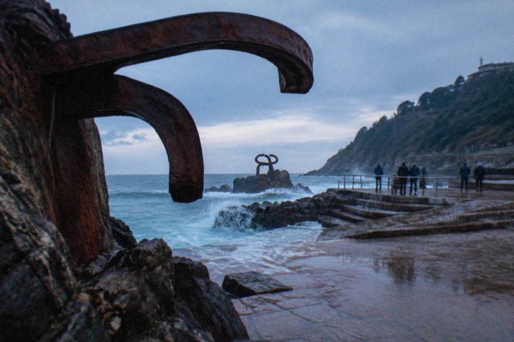 A wide pink granite terrace at the water's edge with curved steel sculptures and a railing, waves washing across the stone