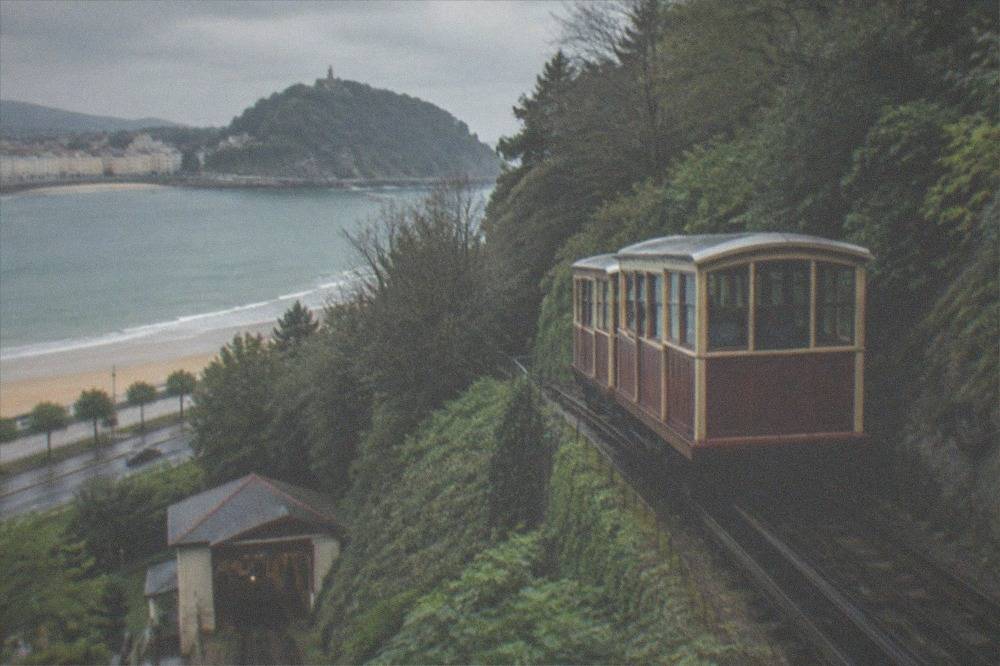 A wooden funicular car on a steep rail track climbing a wooded hill above a beachfront road