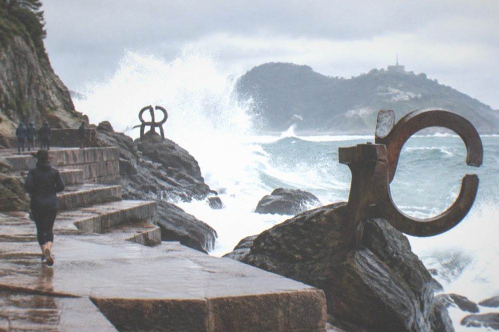 Waves crashing high over dark rocks and steel sculptures, spray bursting upward against a grey sky