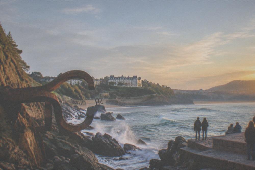 A white palace with gardens sloping to the sea, seen from a promenade beside a wide sandy bay