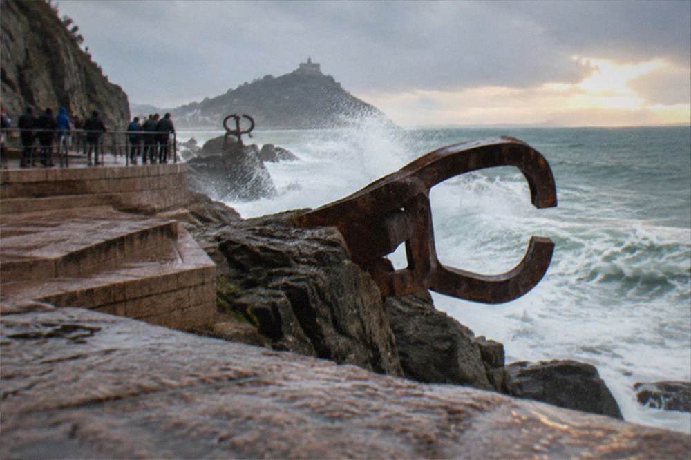People leaning on a railing watching waves explode against steel sculptures on rocks at sunset