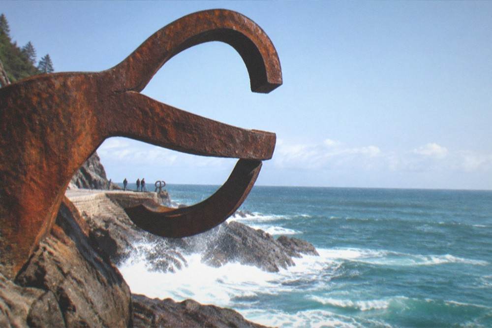 A close view of a rusted steel sculpture shaped like curved fingers, with the sea and horizon beyond it
