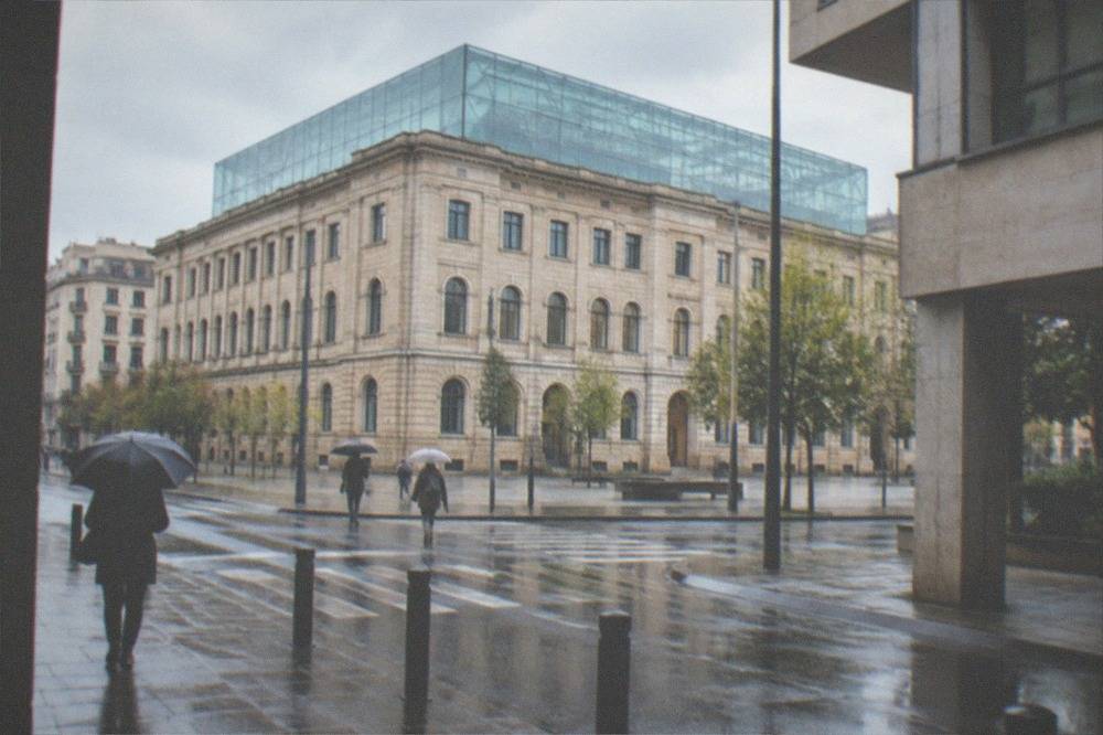 A city street with a modernist building corner and people walking with umbrellas in soft rain