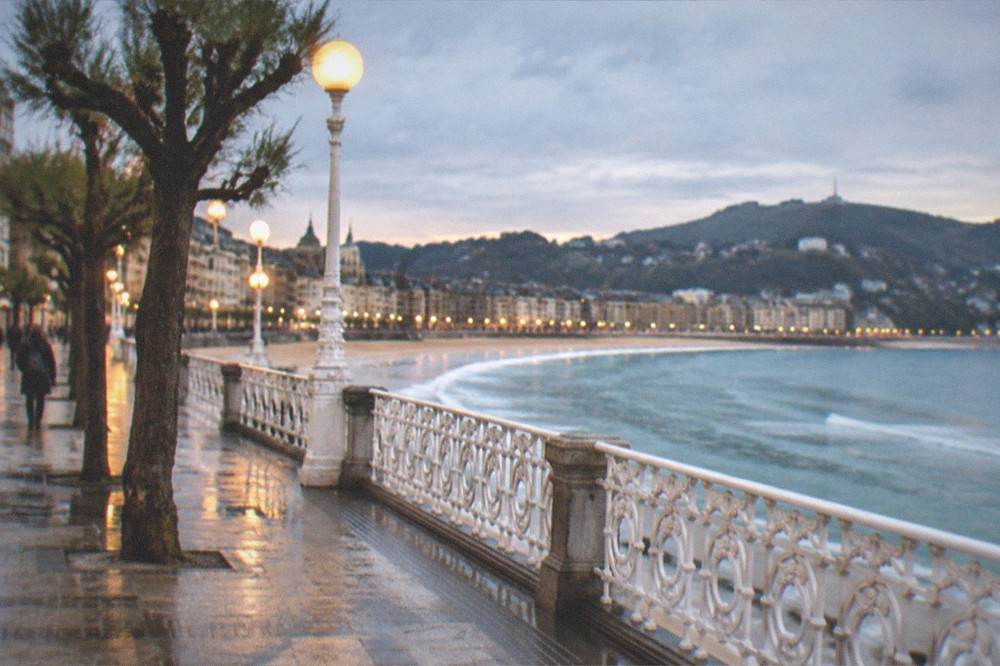 An evening promenade beside a curved city bay, street lamps lit along the railing and hills closing the horizon