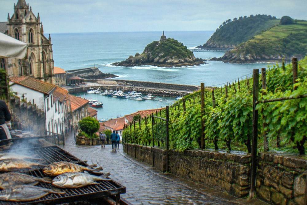 A rocky headland with an iron footbridge reaching out to a small island with a statue, above breaking Atlantic waves