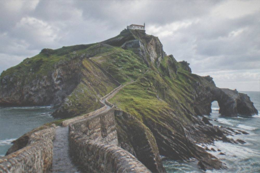 The whitewashed stone hermitage at the top of the islet, seen from the path against an overcast sky