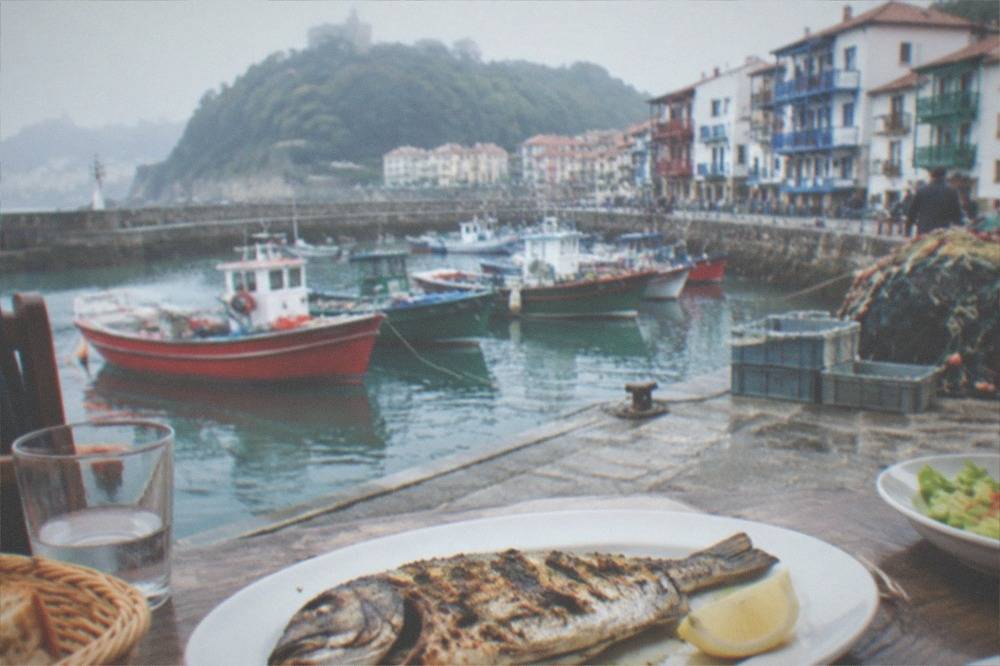 A whole grilled fish being served at a table beside a harbour with fishing boats