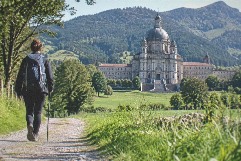 A pilgrim walking a path through green countryside toward the sanctuary complex in the distance