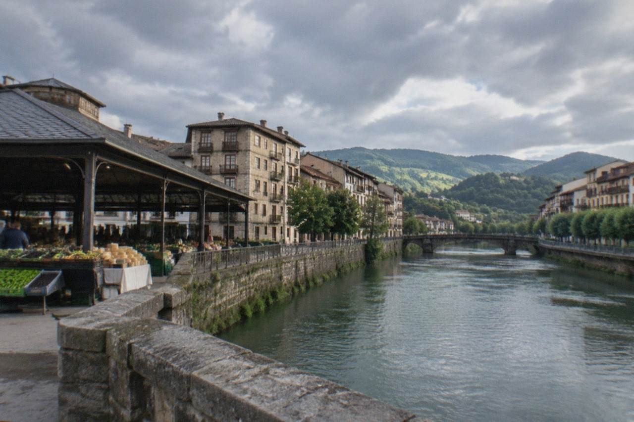 A Basque riverside town seen from a stone bridge with green hills behind it under a cloudy sky