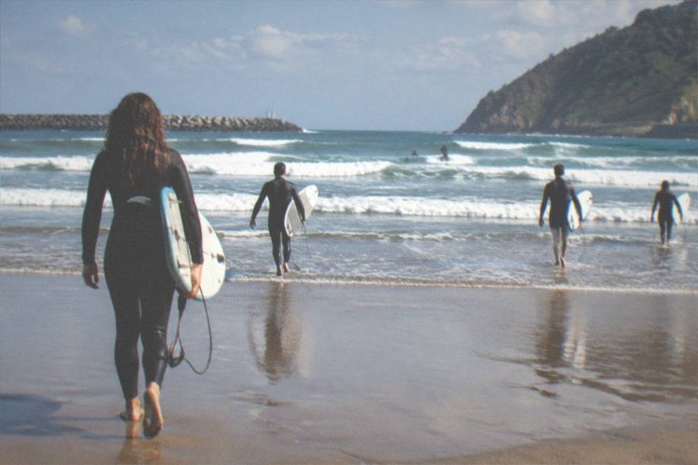 Surfers in wetsuits carrying boards across wet sand toward small waves