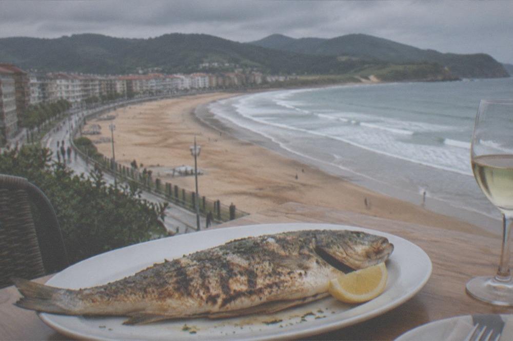 A whole grilled fish on a plate beside a glass of pale white wine on a restaurant table