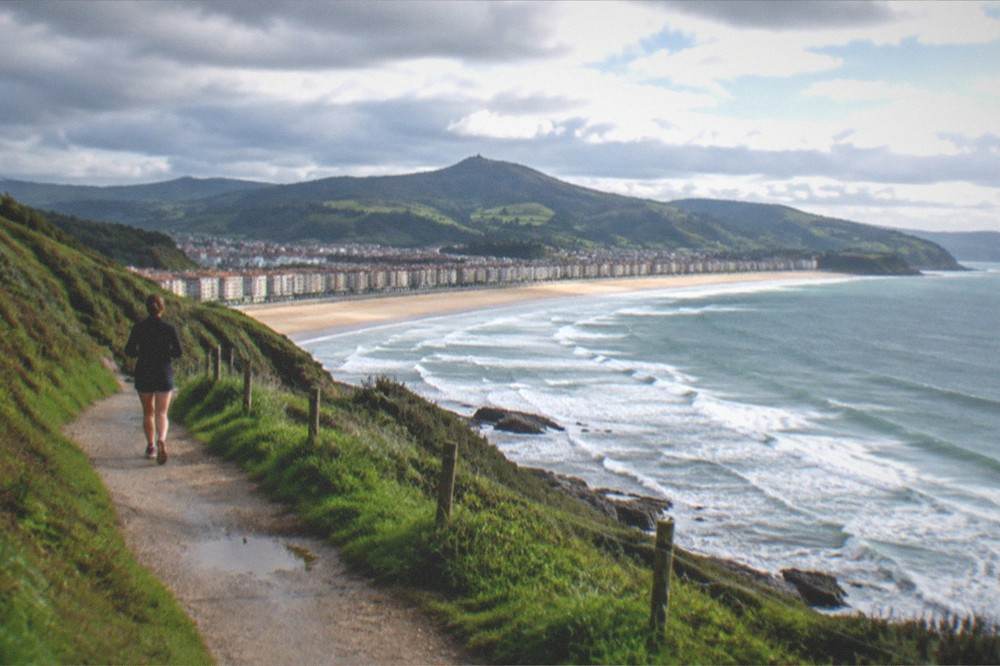 A coastal path on green cliffs with the sea and a distant town below