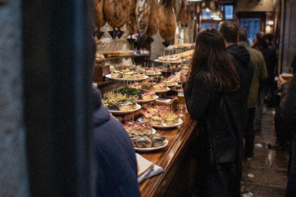 A crowded pintxo bar counter in the Old Town with trays of small skewered portions displayed on the bar top