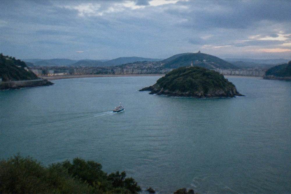 A small boat crossing La Concha bay toward a green island with a lighthouse