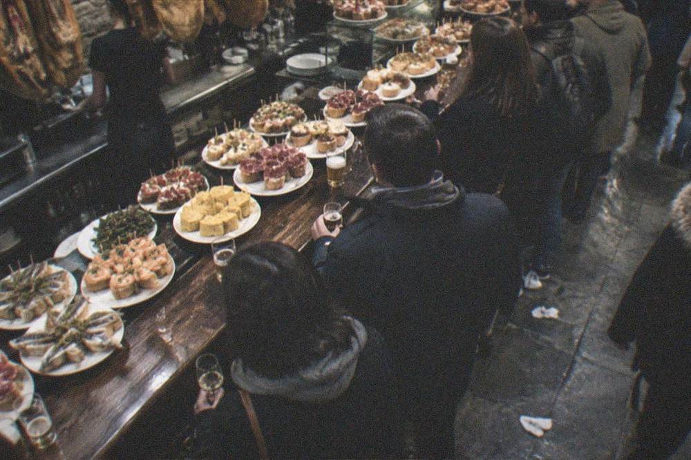 Crowded pintxo bars in an Old Town street at night, with plates of food on the counter