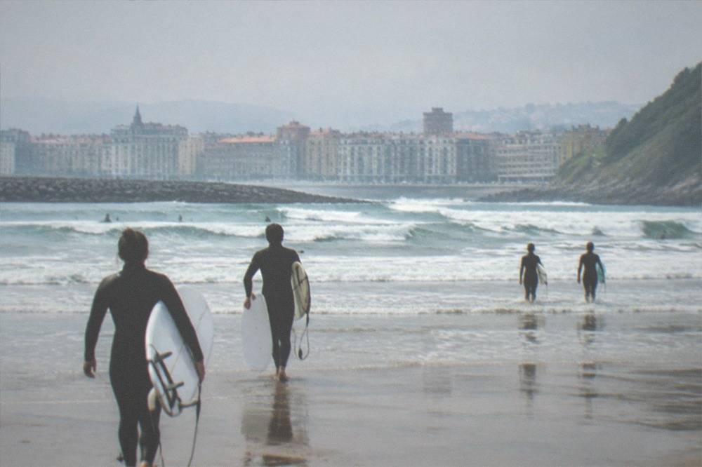 Surfers walking onto Zurriola beach with waves breaking and the Gros rooftops behind