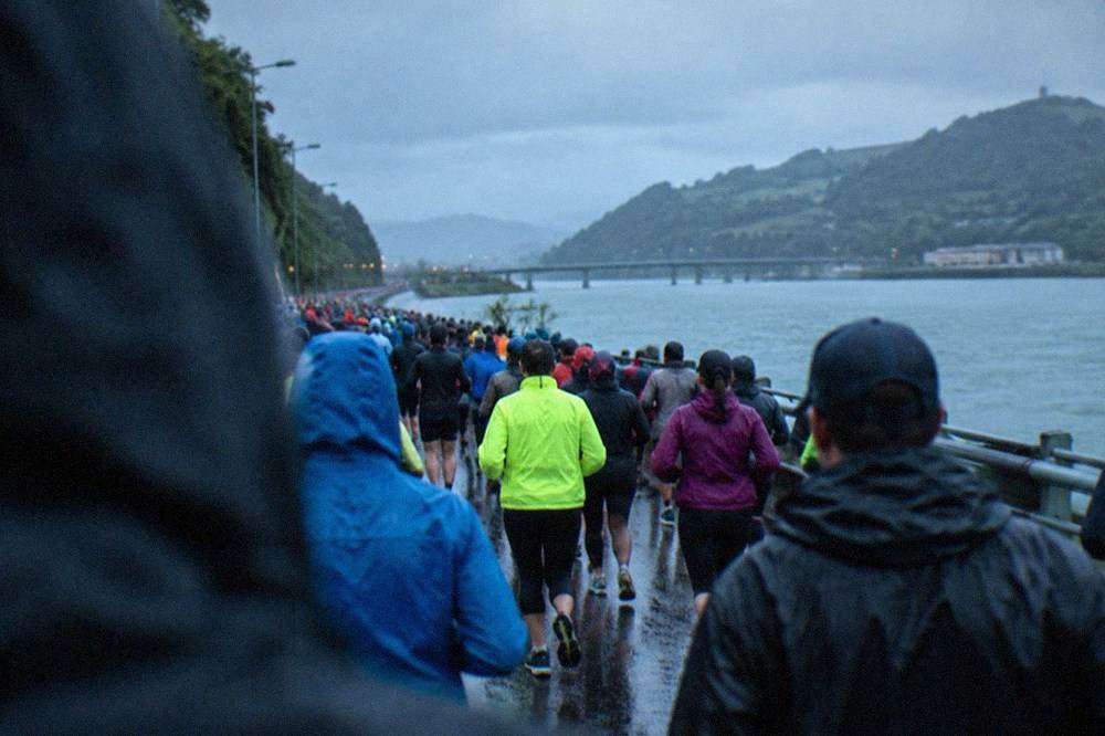 Runners in rain jackets passing a coastal road with green hills