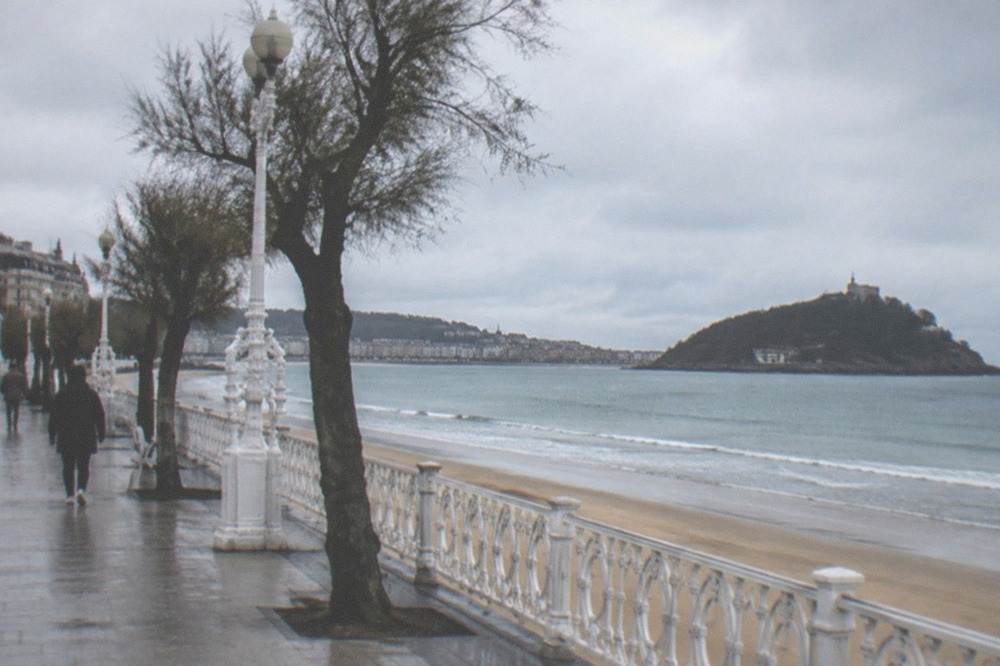 A seafront promenade under grey skies with a wide sandy bay and an island