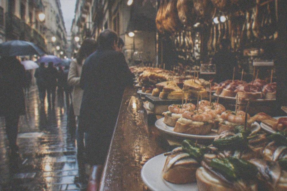 People with umbrellas walking between lit pintxo bars on a rainy evening