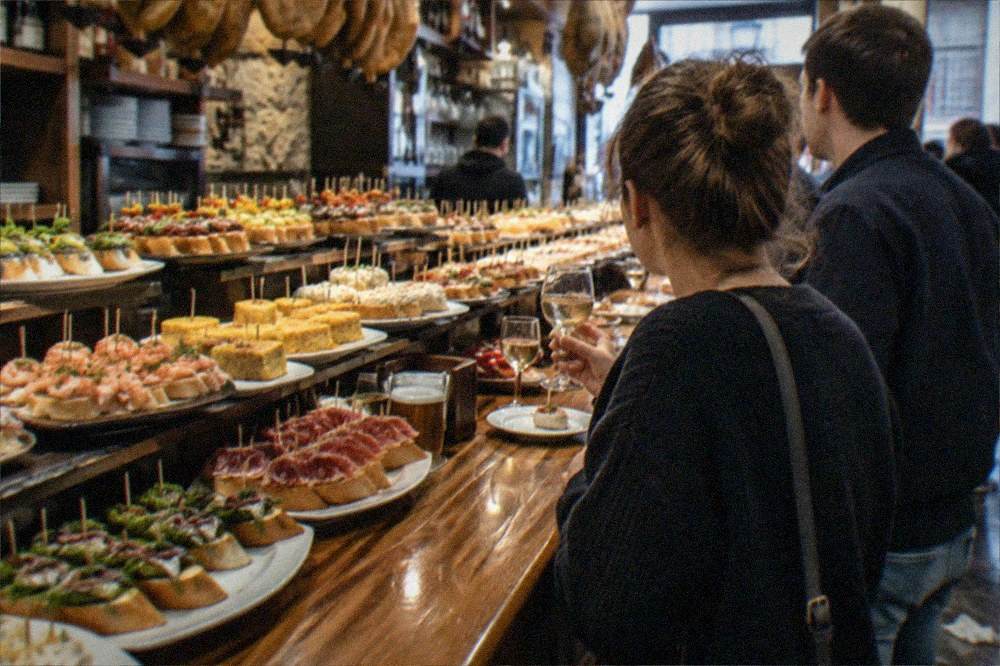 Two people standing at a lit bar counter with small plates and glasses of wine