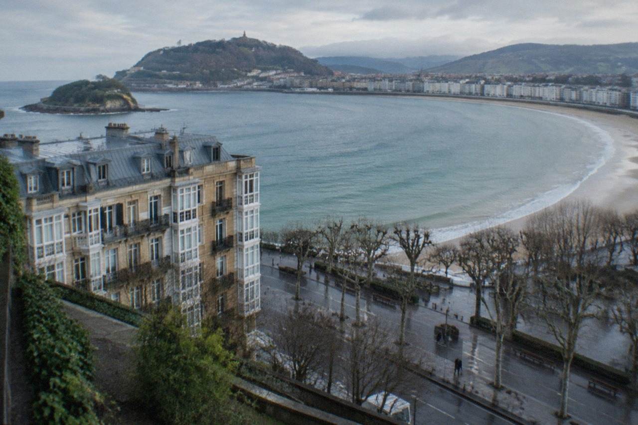 The seafront of San Sebastián seen from above, with La Concha bay curving between green hills and a row of white buildings