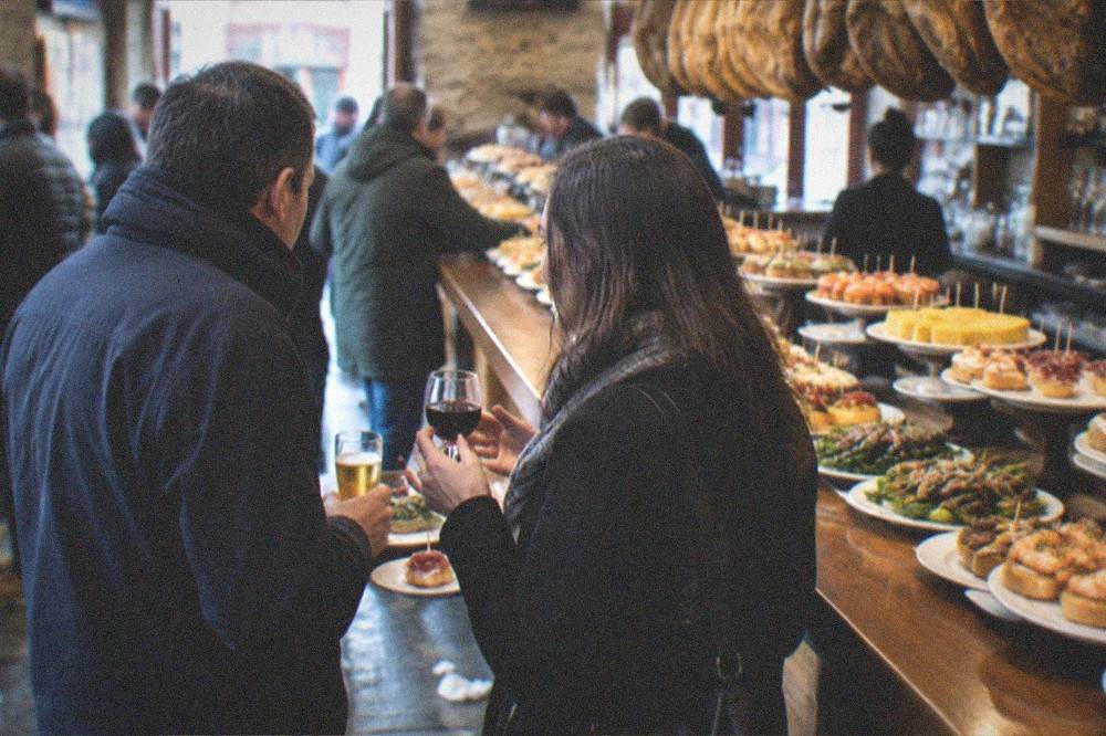 Two people standing at a bar counter with small plates and glasses, mid-conversation