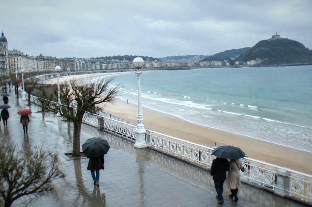 People walking with umbrellas along a wet promenade beside La Concha beach