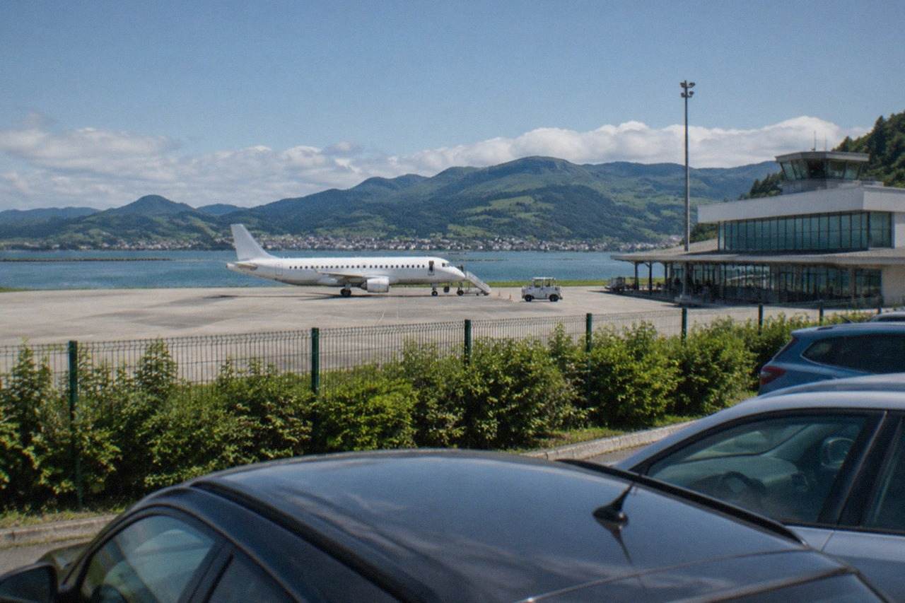 A small regional airport terminal beside green hills with parked cars in the foreground