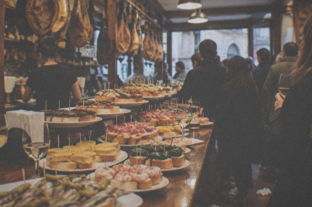 A crowded pintxo bar counter with small plates and glasses of white wine