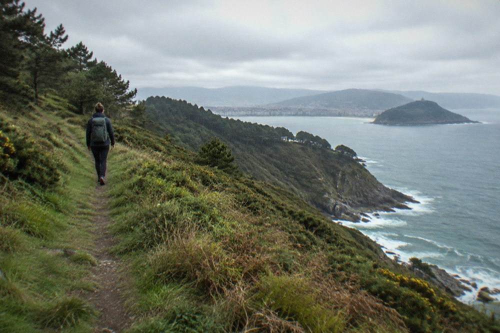 Walker with a backpack on a grassy ridge path above a steep green coastline
