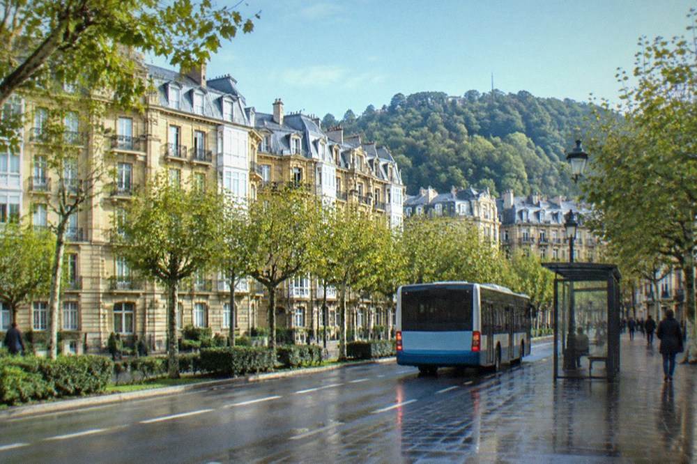 A city bus pulling away from a stop on a tree-lined street in San Sebastián
